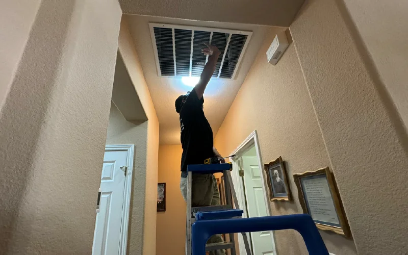 Air duct cleaning technician servicing a ceiling return vent with professional equipment in a residential hallway.